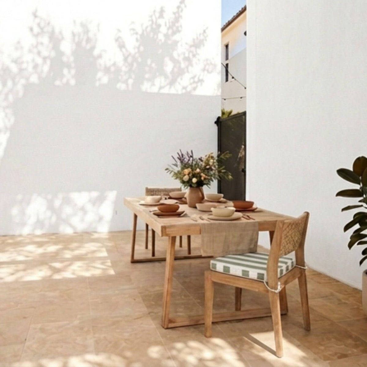 A light natural wood dining table set with a woven-back chair featuring a green and white striped chair cushion, styled with a floral centerpiece and terracotta bowls in a bright, sunlit outdoor patio setting.