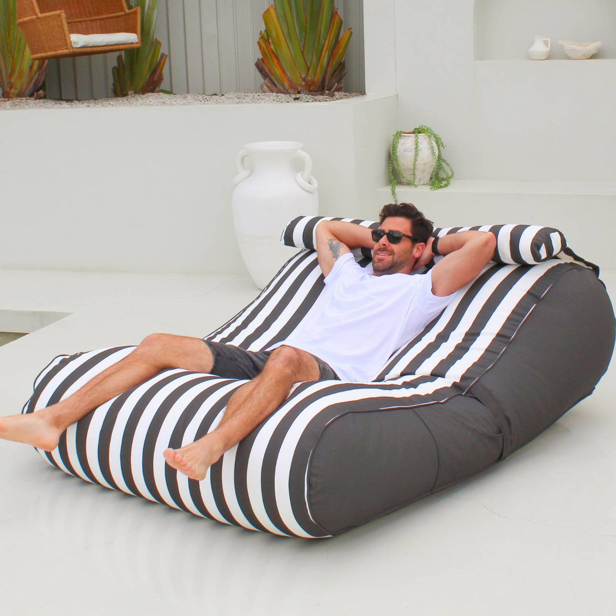 Man stretching out on an Oyster Black Nevaeh double outdoor bean bag sunbed, showing its striped seat and plain side panels, MOSTNEVBB-OYSBLA