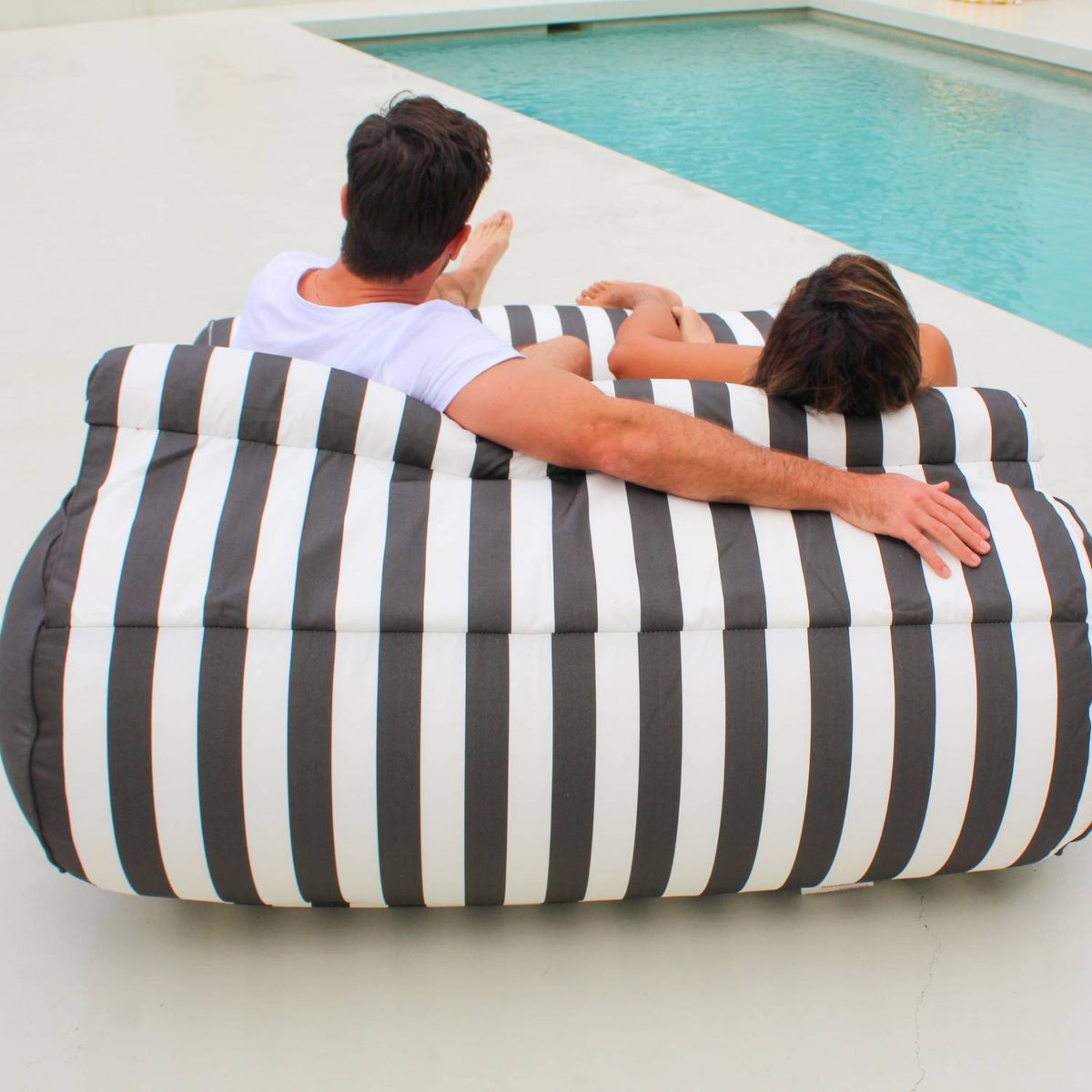 Rear view of a couple sharing an Oyster Black Nevaeh double outdoor bean bag sunbed facing a pool, showing its striped back and bolster cushion, MOSTNEVBB-OYSBLA