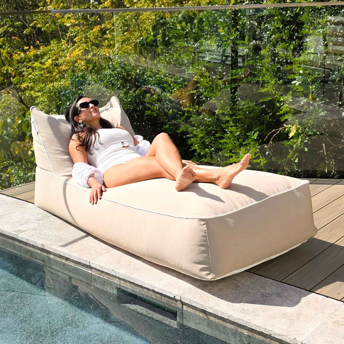 Woman reclining on a Taupe Talia bean bag sunlounger beside the pool, showing its raised backrest and white piping, MOTALFLTBB- TAUP
