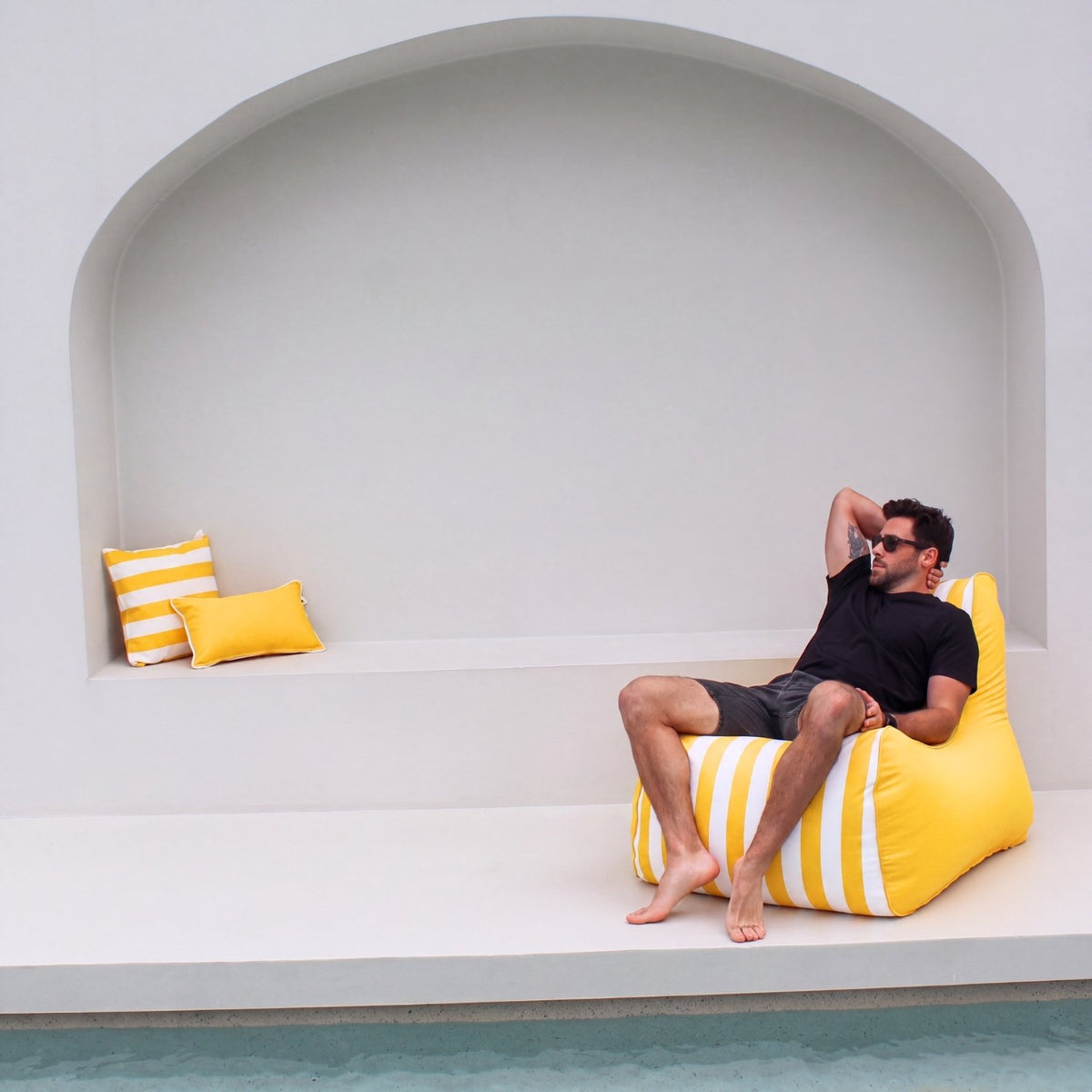 A man wearing sunglasses and a black t-shirt relaxes on a bright yellow and white striped Bora bean bag chair next to a pool, with a matching yellow and white striped pillow on a white bench in the background, MOBORABB-STRI-YEL