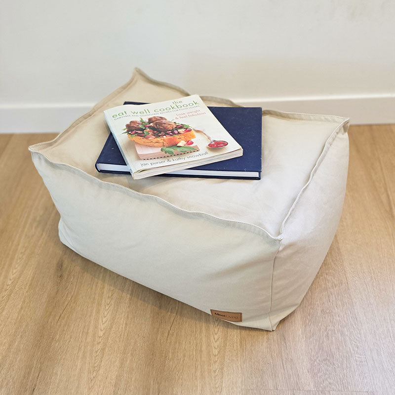 Mooi Living Boss Ottoman in Natural cotton canvas styled alone on timber flooring, with books resting on top of the ottoman.