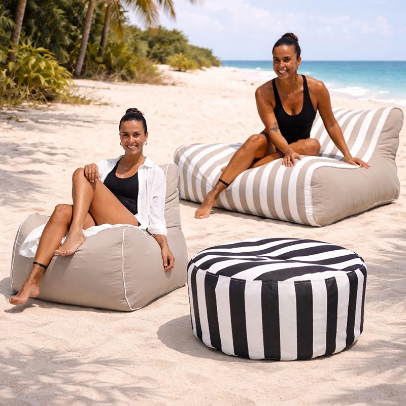 Lifestyle image of the Bora Bora Chair in Taupe and Chill Lounger UV outdoor bean bag by Mooi Living in Taupe Stripe, and Outdoor Ottoman in Oyster Black Stripe with two women sitting on the bean bags on a sandy beach with palm trees and ocean in the background.