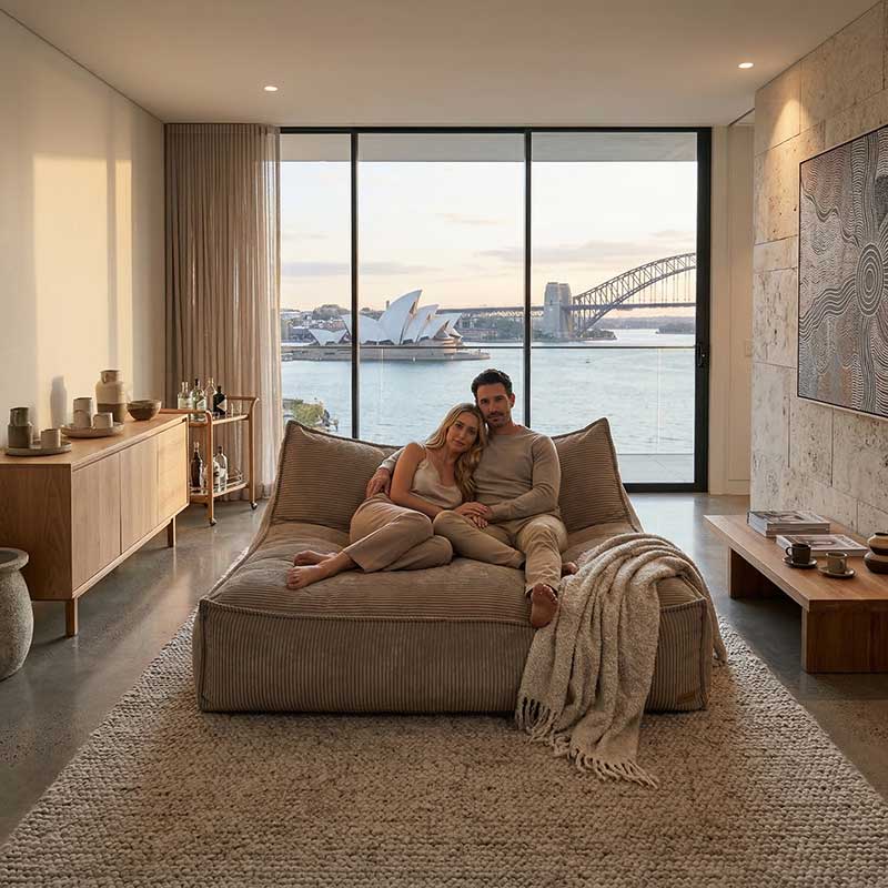 Lifestyle image of the Mooi Double Cord indoor bean bag by Mooi Living in Caramel with a couple sitting on it while relaxing in a hotel room with the beautiful view of the Sydney Opera House in the back.