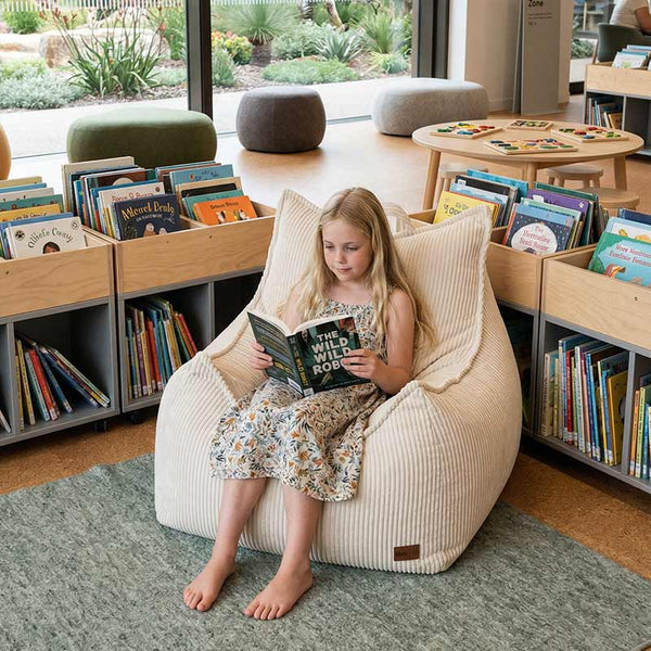 Lifestyle image of the Mini Cord Chair indoor bean bag by Mooi Living in Beige with a child sitting on it while reading a book, in a library setting.