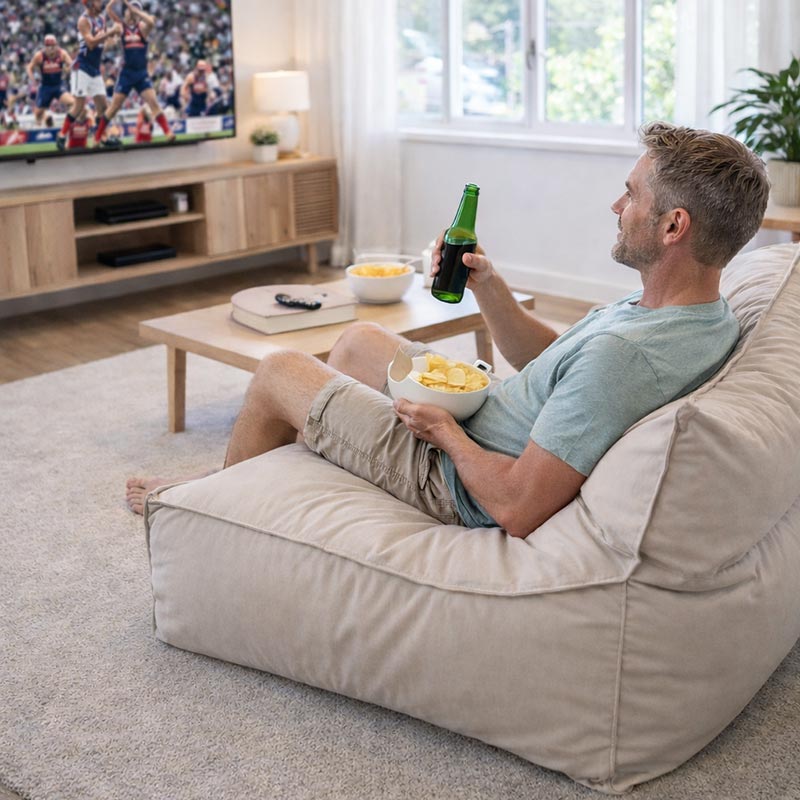 Lifestyle image of Cotton Natural Boss Bean Bag Chair indoor bean bag by Mooi Living, with a man sitting on the bean bag chair in a living room, watching TV and eating snacks.