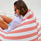 Close-up of a woman reclining on the blush and white striped side of a reversible triangle outdoor bean bag, MOREVSTRIBB-BLUS