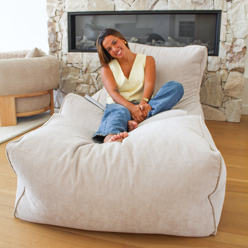 Brooklyn Corduroy bean bag chair in Natural Beige in a modern room with a woman sitting on the bean bag chair in a living room with a stone fireplace.
