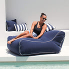 A woman wearing sunglasses sits on a large navy Chill UV outdoor bean bag chair with white piping by Mooi Living, MOCHUVBB-NAVY.
