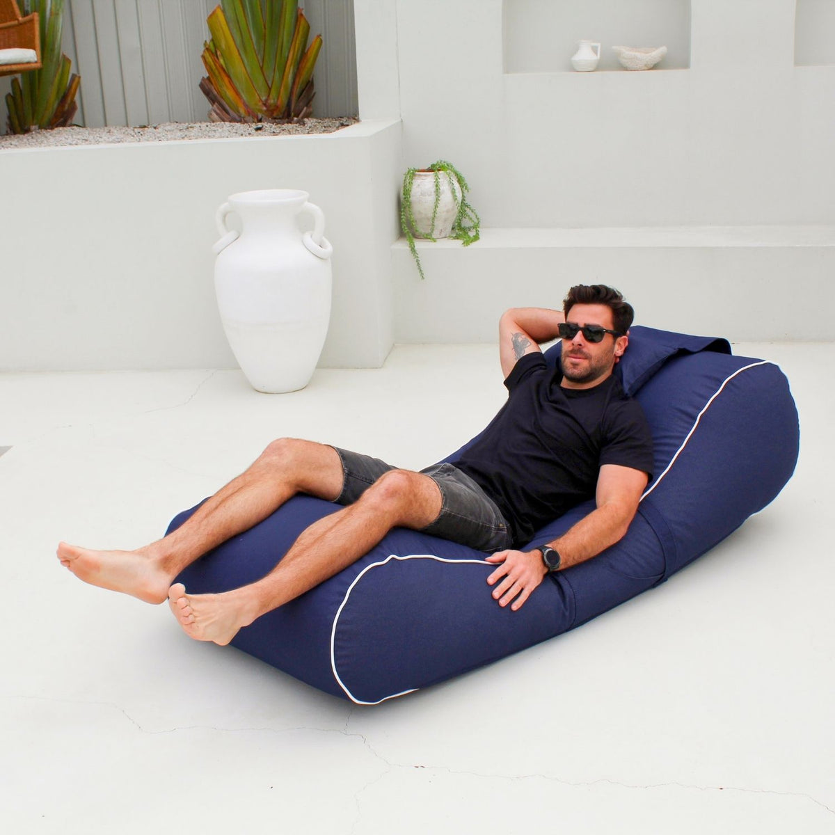 Man reclining on a Mooi Living Navy Isla outdoor bean bag sunlounger with white piping and a matching pillow on a white patio, MOISLABB-NVY
