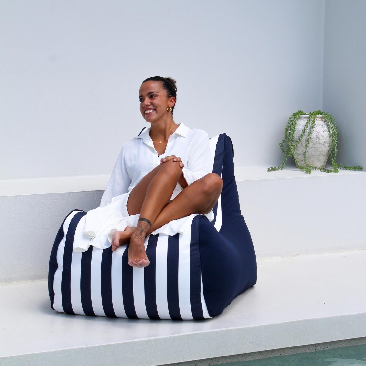 A woman sits cross-legged on a Mooi Living navy and white striped Bora bean bag chair in a bright, modern white outdoor setting, MOBORABB-STRI-NAV