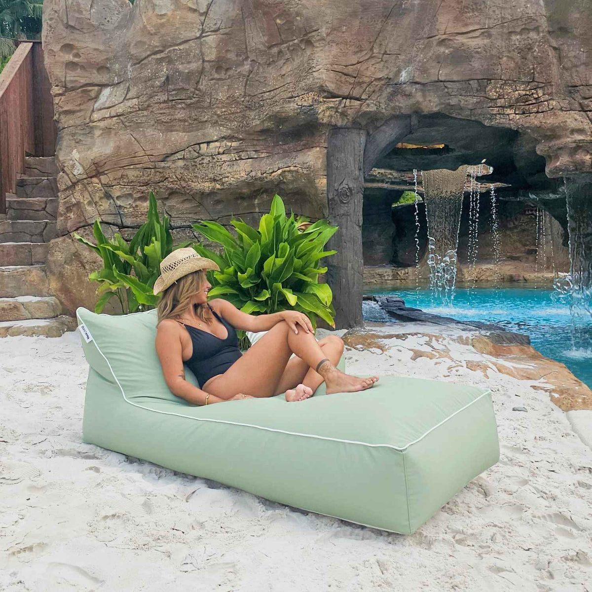 Woman wearing a straw hat relaxing on a Sage Green Talia bean bag sunlounger on sand beside a pool waterfall, MOTALFLTBB-SAGGRE