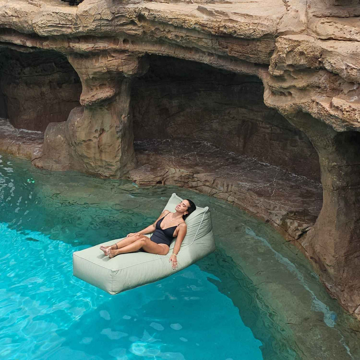 Woman reclining on a Mooi Living Sage Green Talia sunlounger bean bag pool float beside a rock cave, MOTALFLTBB-SAGGRE