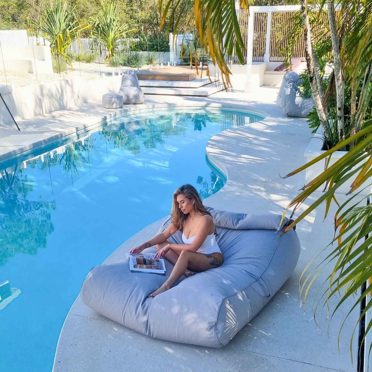Wide view of a woman reading on a Mooi Living Pumice Stone Nevaeh double outdoor bean bag sunbed beside a tropical pool, MOSTNEVBB-PUMSTO