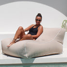 A woman wearing a black swimsuit and sunglasses sits on a large, taupe Chill UV outdoor lounge bean bag chair with white piping, positioned next to a white pool deck under a bright sun, MOCHUVBB-TAUP