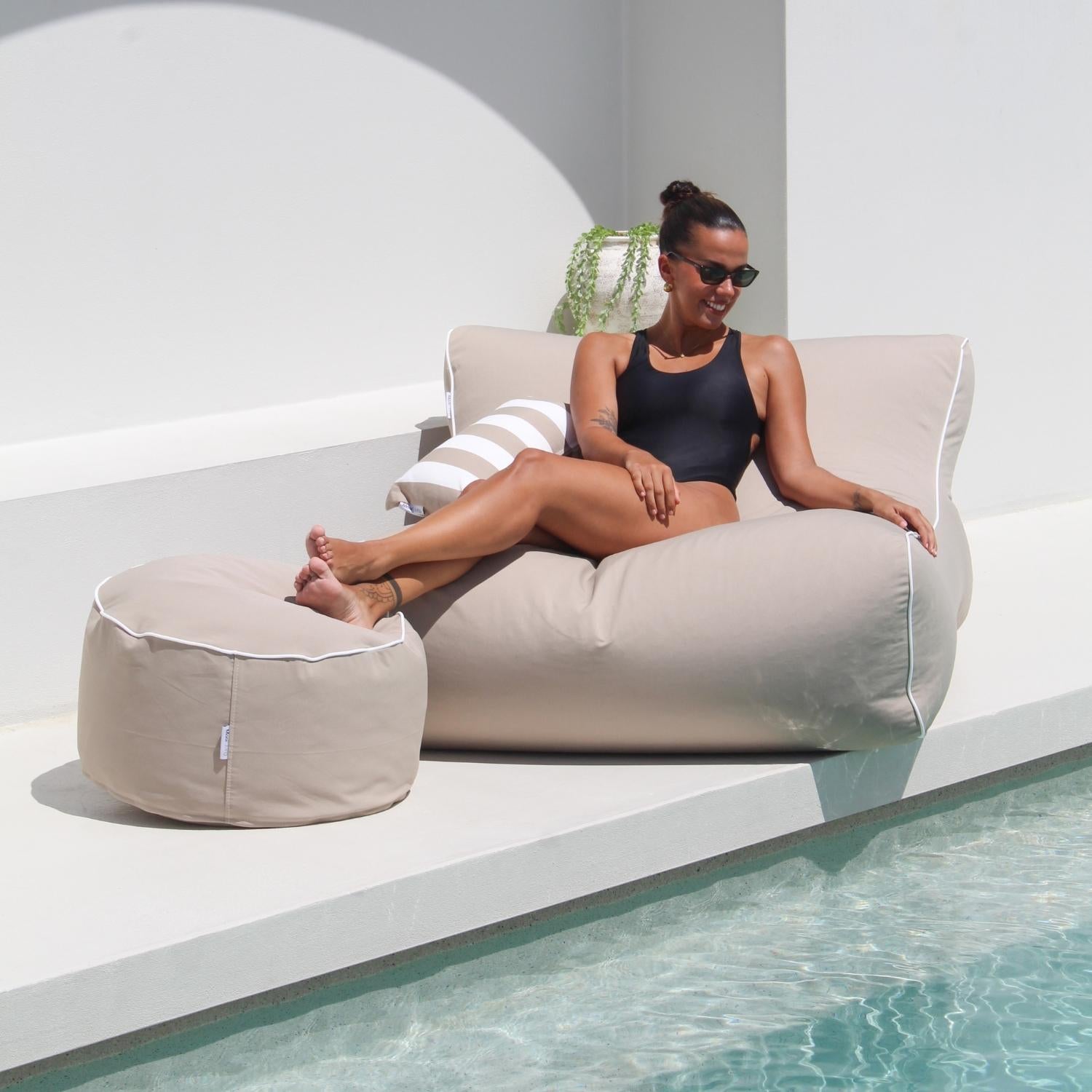 A woman in a black swimsuit and sunglasses relaxes on a taupe Chill UV outdoor lounger next to a pool, with a matching round ottoman and a striped pillow in a modern white patio setting.
