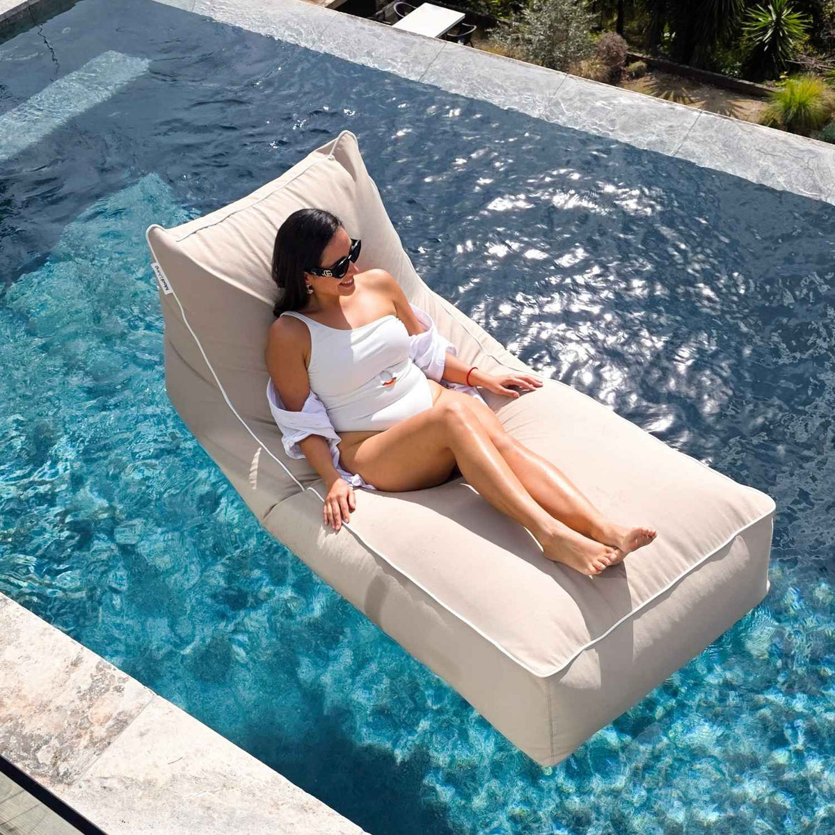 Overhead view of a woman in white swimwear reclining on a Mooi Living Taupe Talia bean bag pool float, MOTALFLTBB- TAUP