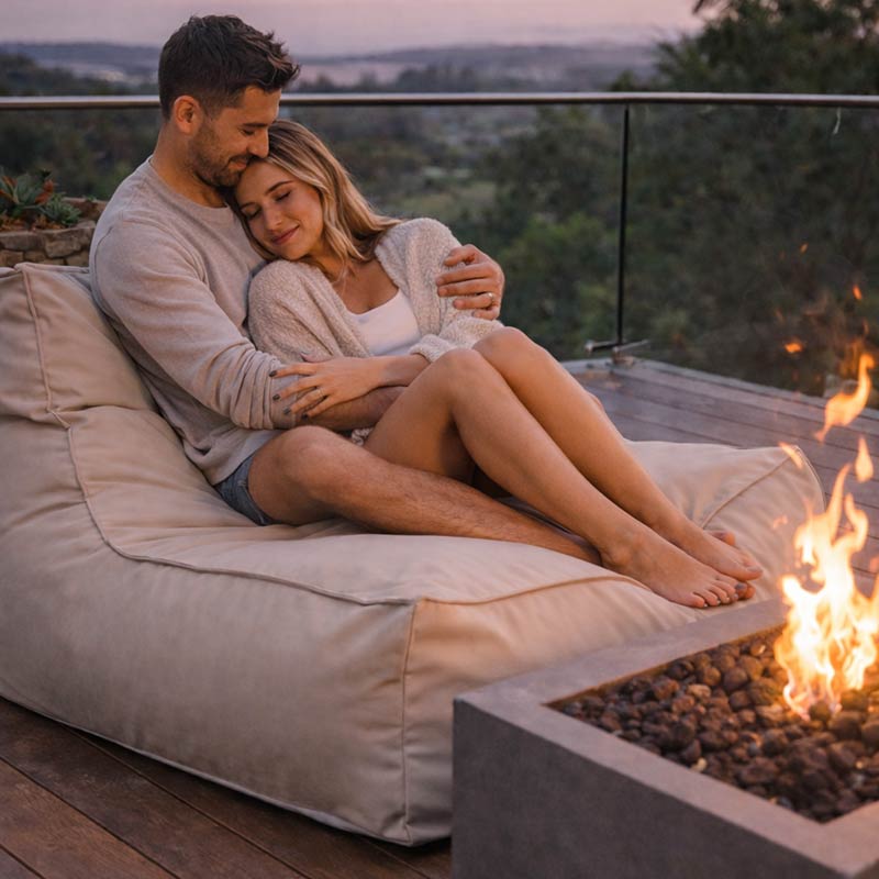Lifestyle Image of the Natural Beige Lana Lounger outdoor bean bag by Mooi Living with a couple sitting on the lounger bean bag by a fire pit with a scenic background