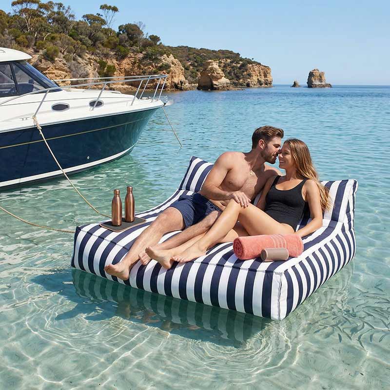 Lifestyle Image of the Navy Stripe Lana Lounger outdoor bean bag by Mooi Living floating in the water, tied to a boat with two models lounging comfortably. 