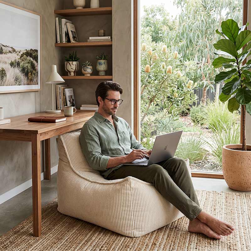Lifestyle image of the Cord Chair indoor bean bag by Mooi Living in Beige with a man sitting on the bean bag while using a laptop in a home office with plants and a desk.