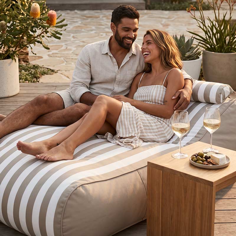 Couple sitting on a striped Nevaeh Sunbed outdoor bean bag by Mooi Living with wine glasses and snacks, surrounded by plants.