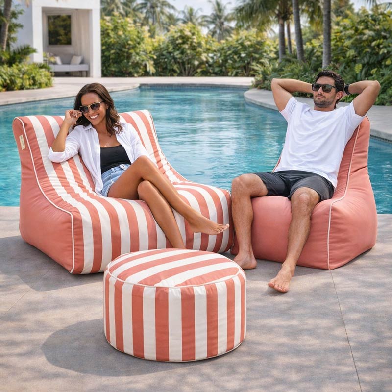 Lifestyle image of the Chill Lounger UV in Blush Stripe, Bora Bora Chair, outdoor bean bag by Mooi Living in Blush, and Outdoor Ottoman in Blush Stripe with two people sitting on the bean bags by a poolside. Perfect for poolside relaxation.